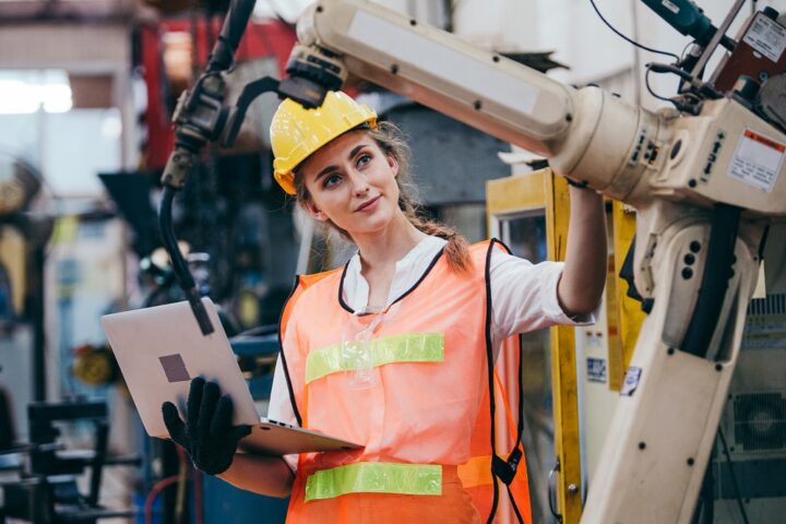 Industrielle Ingenieurin oder Technikerin mit Schutzhelm und Uniform, die einen Laptop benutzt und an einer Roboterarmmaschine arbeitet.