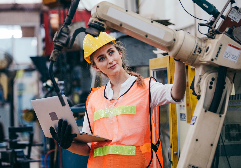 Industrielle Ingenieurin oder Technikerin mit Schutzhelm und Uniform, die einen Laptop benutzt und an einer Roboterarmmaschine arbeitet.