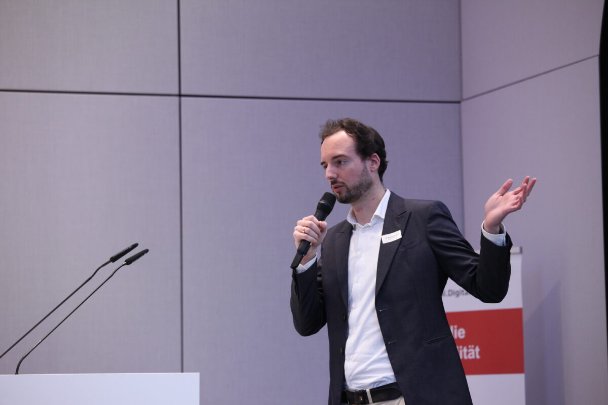 Maximilian Bock steht während seines Vortrags auf der Bühne und präsentiert mit einem Mikro in der Hand.