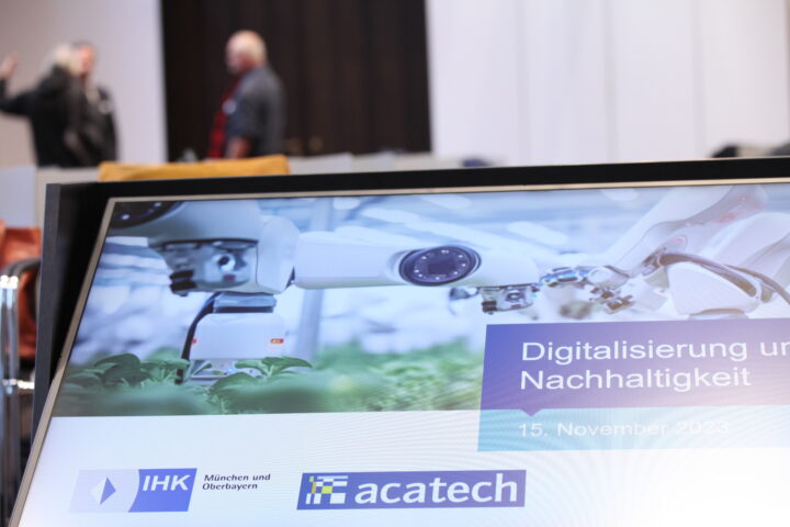 Screen zeigt den Titel der Veranstaltung "Digitalisierung und Nachhaltigkeit". Im Hintergrund tauschen sich Personen zu dem Thema aus.